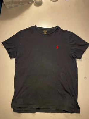 Svart t-shirt från Polo Ralph Lauren - Klassisk svart t-shirt från Polo Ralph Lauren med röd broderad logga på bröstet. Tillverkad i 100% bomull och har en normal passform med korta ärmar. Perfekt basplagg för en stilren look.
