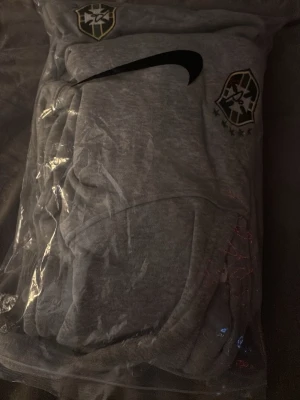 Grå Nike hoodie Brasilien - Snygg grå hoodie från Nike med Brasilien-emblem och svart swoosh på bröstet. Hoodien har huva och är tillverkad i mjukt material, perfekt för en avslappnad och sportig stil. Passar dig som gillar fotboll och streetwear.