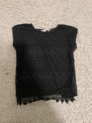 Svart spetsblus med ärmlös design - Svart blus med spets framtill och dekorativ kant nertill. Ärmlös modell med rund halsringning och mjukt tyg på baksidan. Perfekt för dig som gillar detaljer och en stilren look.