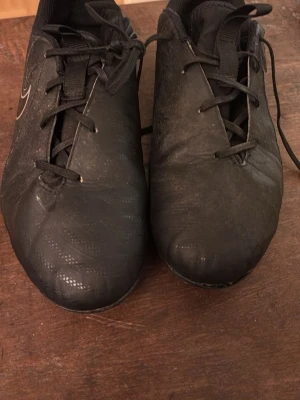 Svarta Nike fotbollsskor - Säljer ett par svarta fotbollsskor från Nike med klassisk vit swoosh på sidan. Skorna har snörning och är tillverkade i syntetmaterial för bra grepp och kontroll på planen. Perfekta för dig som vill ha stilrena och funktionella skor till match eller träning.