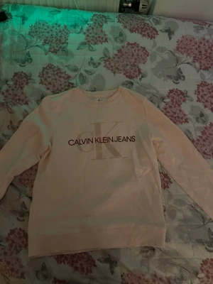Ljusrosa sweatshirt från Calvin Klein strl 14 years - Ljusrosa sweatshirt från Calvin Klein Jeans med stort CK-tryck och logga framtill. Tröjan har rund halsringning, långa ärmar och ribbade muddar. Perfekt för en avslappnad och trendig look.