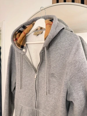 Mc cardigan bberry - Säljer en grå hoodie från Burberry med dragkedja och klassiskt rutigt foder i huvan. Hoodien har fickor framtill och justerbar dragsko i huvan. Perfekt för dig som gillar stilrena och exklusiva plagg.