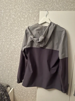 Grå och mörkgrå vindjacka Under Armour - Snygg tvåfärgad vindjacka från Under Armour i grått och mörkgrått. Jackan har huva, dragkedja framtill och praktiska fickor. Tillverkad i lätt och tåligt syntetmaterial, perfekt för blåsiga dagar.