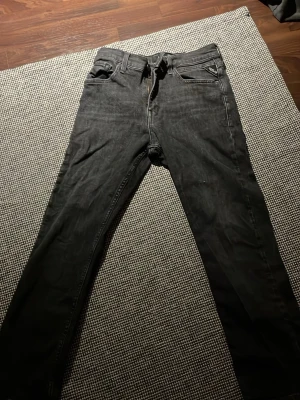Svarta Replay jeans med smal passform - Säljer ett par mörk grå jeans från Replay med otroligt bra passform och klassisk femficksdesign. Jeansen har dragkedjegylf, snygga detaljer vid fickorna och Replay-logga på bakfickan. Perfekta för dig som gillar en stilren och modern look.