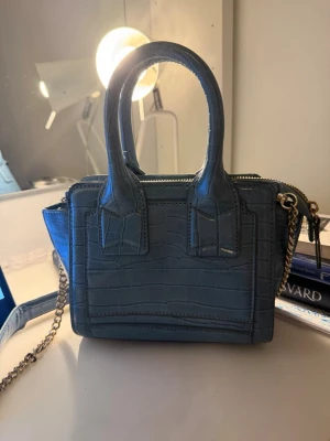 Blå minihandväska med kedja - Snygg blå minihandväska i croco-mönstrat konstläder med två bumper handtag och guldfärgad kedjerem. Väskan har dragkedja upptill och en stilren, boxig form som ger en trendig look. Skriv ifall ni vill ha måtten