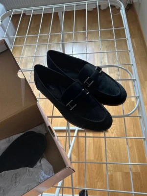 Nya svarta loafers med metallspänne storlek 38 - Stilrena svarta loafers från Graceland i mockaimitation med snygga silverfärgade metallspännen framtill. Klassisk design med rundad tå och låg sula, perfekta för en trendig och avslappnad look. Slutsålda vid inköp.