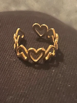 Stål Guldfärgad ring med hjärtan - Säljer en snygg guldfärgad ring med öppet mönster av flera sammankopplade hjärtan runt hela ringen. Ringen har en modern och lekfull design som sticker ut och passar perfekt för dig som gillar unika accessoarer. Materialet ser ut som metall med guldfinish.