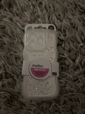 Mobello Wavy Glitter mobilskal i vitt - Snyggt mobilskal från Mobello med vågig form och glitterdetaljer. Passar iPhone och har extra skydd runt kameran. Perfekt för dig som vill ha ett skal som sticker ut. Aldrig använt och säljer då jag ångar mitt köp💕PASSAR IPHONE 14PRO