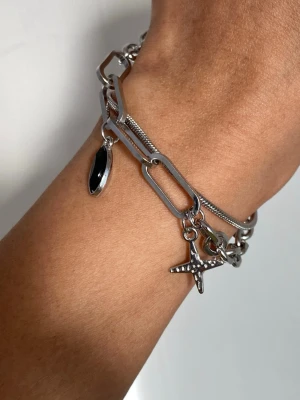 Silverfärgat kedjearmband med berlocker - Armband i silverfärgad metall med stora kedjelänkar och två berlocker, en svart oval och ett kors. Justerbar längd med extra kedja och platt bricka. Snyggt och trendigt för dig som gillar detaljer.