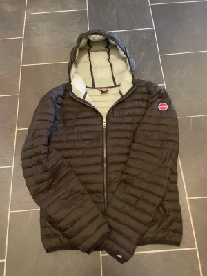 Svart dunjacka med huva från Colmar - Snygg svart dunjacka från Colmar med quiltad design och huva. Jackan har dragkedja framtill, två sidofickor och ett Colmar-märke på ärmen. Insidan är ljusgrå vilket ger en schysst kontrast. Perfekt för kalla vinterdagar. Har två lappade hål på armen.