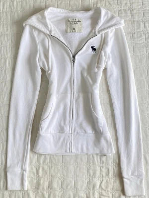 Vit hoodie från Abercrombie & Fitch - Snygg vit hoodie med dragkedja från Abercrombie & Fitch. Klassisk passform med huva, dragsko och två fickor framtill. Liten mörkblå logga med älg på bröstet. Perfekt för en avslappnad och stilren look.
