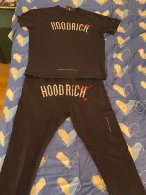 Svarta mjukisbyxor från Hoodrich - Säljer ett par svarta mjukisbyxor från Hoodrich med stor vit logga och röd detalj framtill. Byxorna har en avslappnad passform och en ficka på sidan med dragkedja. Perfekta för chill eller streetwear-stil. Tröjan är M och byxorna L