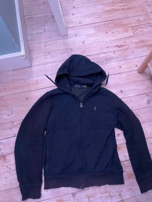 Mörkblå cardigan från polo - Mörkblå hoodiejacka från Polo Ralph Lauren med dragkedja framtill och broderad logga på bröstet. Jackan har huva med snörning och är tillverkad i ett mjukt material. Perfekt för en avslappnad och stilren look.