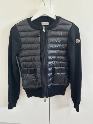 Svart cardiganjacka från Moncler - Snygg svart cardiganjacka från Moncler med quiltad framsida i glansigt material och stickade ärmar. Jackan har två fickor framtill med knappar, dragkedja och Moncler-logga på ärmen. Perfekt för dig som vill ha en lyxig stil.
