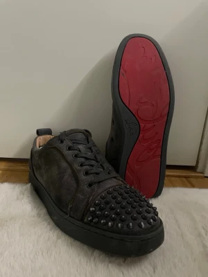 Christian Louboutin (äkta) - Unika svarta sneakers från Christian Louboutin med mattsvarta nitar på tån och klassisk röd sula. Skorna har en låg profil, svarta skosnören och är tillverkade i mocka. Perfekta för dig som vill sticka ut med en edgy och lyxig stil.
