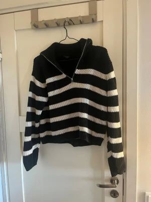 Svartvitrandig stickad tröja Lindex - Stickad tröja från Lindex i svart med breda vita ränder. Tröjan har halv dragkedja framtill, hög krage och ribbstickade muddar. Perfekt för dig som gillar en avslappnad men trendig look.