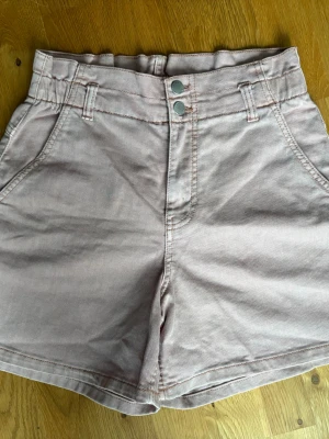 Ljusrosa högmidjade shorts - Snygga ljusrosa shorts med hög midja och dubbla knappar framtill. Shortsen har bälteshällor, två fickor fram och två bakfickor. Perfekta för varma dagar och lätta att matcha med olika toppar.