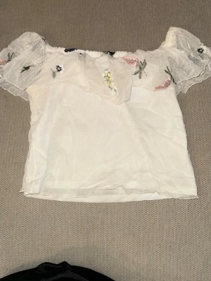 Vit offshoulder blus med broderi - Söt vit offshoulder blus med volang och broderade blommor i olika färger. Tunn och luftig med korta ärmar och romantisk känsla. Perfekt för varma dagar och en trendig look.