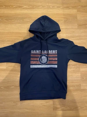 Mörkblå hoodie från Saint Laurent - Mörkblå hoodie från Saint Laurent med tryck framtill i vitt och rött samt texten 'SAINT LAURENT'. Klassisk känguruficka och justerbar huva. Tillverkad i mjukt material som känns skönt mot huden. Perfekt för en avslappnad och trendig look.