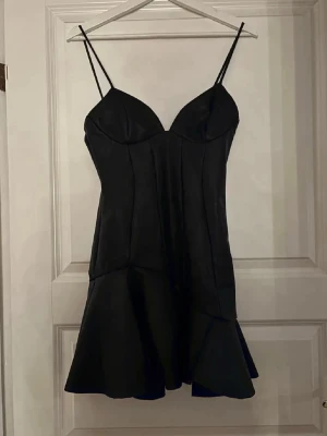 Svart kort klänning med volang - Snygg svart kort klänning med smala axelband och markerad byst. Klänningen har en figurnära passform upptill och volang nertill som ger en cool och trendig look. Perfekt för dig som vill sticka ut med en stilren och elegant outfit. Aldrig använd!! 