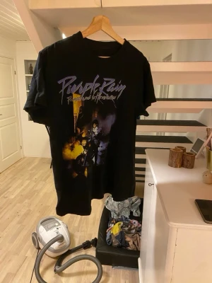 Svart Purple Rain t-shirt - Säljer en svart t-shirt med ikoniskt Purple Rain-tryck framtill i lila och gult. T-shirten har rund hals och korta ärmar. Perfekt för dig som gillar musik och vill ha en snygg statement-tee i garderoben.