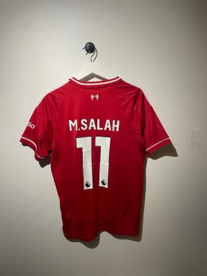 Liverpool fotbollströja Salah 11 Adidas - Röd Liverpool matchtröja från Adidas med vita detaljer och tryck. Framsidan har klubbmärke och sponsorlogga, baksidan har M. Salah och nummer 11. Korta ärmar och sportig passform, perfekt för fotbollsfans.