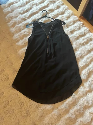 Svart ärmlös linnetopp med dragkedja - Snygg svart ärmlös linnetopp  med en dekorativ guldfärgad dragkedja framtill. Toppen har en lätt rundad nederkant och är tillverkad i ett tunt, mjukt material som känns skönt mot huden. Perfekt att styla med jeans eller kjol.