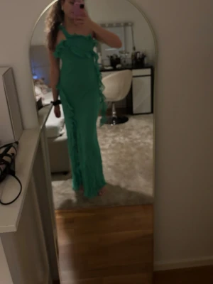 Grön långklänning med volanger - Snygg grön långklänning med volangdetaljer och axelbandslös design. Klänningen har en figurnära passform och är perfekt för dig som vill sticka ut. Materialet känns lätt och luftigt, vilket gör den bekväm att bära under varma dagar.