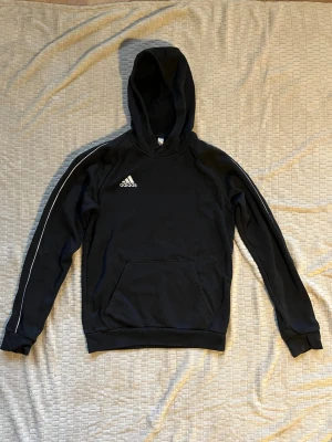 Svart hoodie från Adidas - Snygg svart hoodie från Adidas med vit logga på bröstet och vita ränder längs ärmarna. Klassisk känguruficka framtill och huva. Tillverkad i mjukt material som känns skönt mot huden. Mått: Längd 69cm, Bredd under armar 52cm, Bredd nertill 42cm. Material: 70% bomull, 30% polyester.
