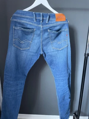Blå jeans från Replay med raka ben - Säljer ett par klassiska blå jeans från Replay med raka ben och snygga detaljer på bakfickorna. Jeansen har fem fickor, ljus tvätt och läderpatch med logga i midjan. Perfekta för en avslappnad och trendig stil.