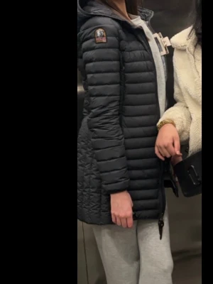 Svart lång dunjacka Parajumpers - Snygg svart lång dunjacka från Parajumpers med huva och dragkedja framtill. Jackan har quiltad design, smal passform och logotyp på ärmen. 