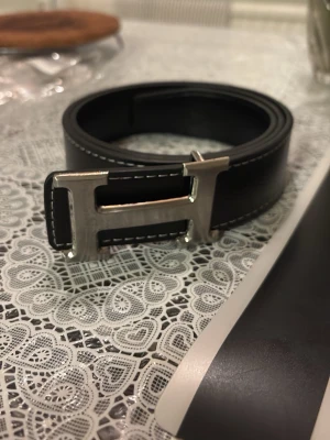 Svart märkesbälte - Snyggt svart märkesbälte med ett stort H-format metallspänne i silver. Skärpet har vita kontrastsömmar längs kanterna och är gjort i slätt skinn. Perfekt accessoar för att lyfta vilken outfit som helst.