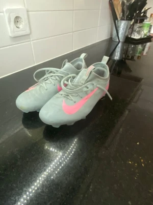 Nike grå-rosa fotbollsskor - Säljer ett par gråa fotbollsskor från Nike med en stor rosa swoosh på sidan och rosa detaljer på insidan. Skorna har snörning och dragflik baktill för enkel påtagning. Perfekta för dig som vill sticka ut på planen!