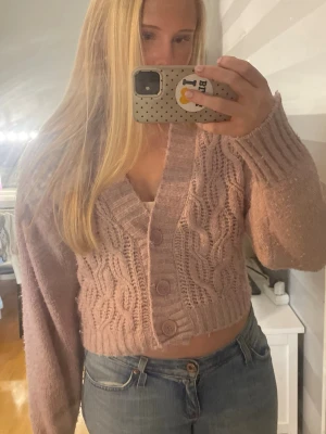 Rosa kabelstickad cropped kofta - Säljer en rosa kabelstickad cropped kofta med tre knappar framtill. Koftan har v-ringning, långa ärmar och ribbade muddar. Perfekt att matcha med jeans för en avslappnad look.