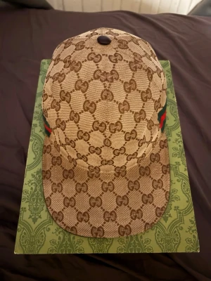Gucci keps med GG-mönster - Snygg beige keps från Gucci med klassiskt GG-monogram över hela kepsen. Har gröna och röda ränder på sidan som ger den en lyxig touch. Kepsen har böjd skärm och är tillverkad i canvasmaterial.