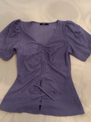 Lila topp med puffärm från BikBok - Superfin lila topp från BikBok med puffärmar och v-ringning. Toppen har snörning framtill som ger en snygg detalj och markerar midjan. Materialet känns mjukt och stretchigt, perfekt till alla tillfällen när du vill känna dig extra söt.