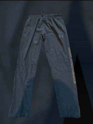 Svarta mjukisbyxor från Under Armour - Säljer ett par svarta mjukisbyxor från Under Armour med resår i midjan och dragsko. Byxorna har raka ben, sidofickor och en snygg logga samt texten UNDER ARMOUR i reflextryck längs ena benet. Perfekta för chill eller träning.