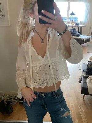 Vit broderad blus med volanger - Söt vit blus med broderade detaljer och volanger. Blusen har v-ringning med knytband och långärmade ärmar med spetskant. Kortare modell som ger en trendig look. Perfekt att matcha med jeans för en avslappnad stil.