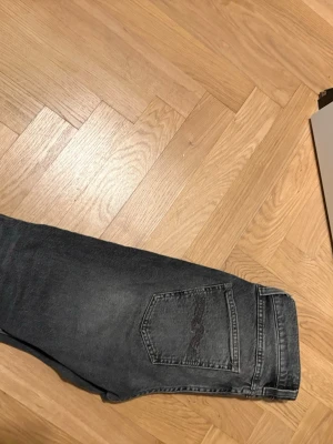 Gråa jeans från Nudie Jeans - Säljer ett par svarta jeans från Nudie Jeans med klassisk femficksdesign och raka ben. Jeansen har en snygg tvättad look och detaljerade sömmar på bakfickorna. Perfekta för dig som gillar stilrena och tidlösa jeans. Tvättade skulle säga att dem sitter i 27