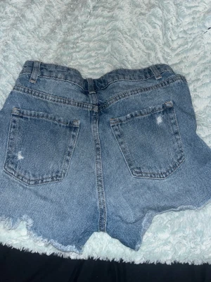 Blå jeansshorts med slitningar - Säljer ett par blå jeansshorts med rå kant och slitna detaljer både fram och bak. Shortsen har två fickor, bälteshällor och knappgylf. Perfekta för sommaren och ger en avslappnad vibe.