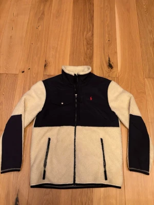 Beige teddyjacka från Polo Ralph Lauren - Säljer nu min helt nya aldrig använda teddyjacka från Polo Ralph Lauren (Endast testad) med mörkblå detaljer på axlar, bröst och armbågar. Jackan har dragkedja framtill, två fickor med dragkedja och en bröstficka med knapp. Mysigt teddyfoder och hög krage för extra värme.