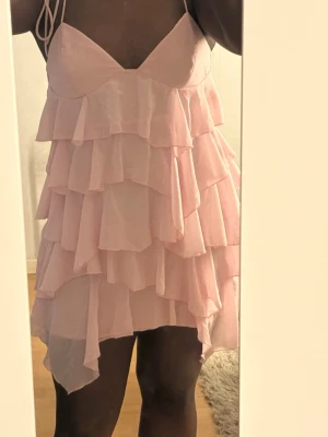 Ljusrosa volangklänning med smala band - Supersöt ljusrosa kortklänning med flera lager volanger och tunna axelband. Klänningen har v-ringad urringning och är tillverkad i ett lätt, fladdrigt material som ger en romantisk känsla. 