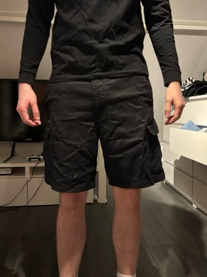 Svarta cargoshorts med fickor - Säljer ett par svarta cargoshorts med flera praktiska sidofickor och klassisk passform. Shortsen har en rak modell och är perfekta för dig som gillar en enkel men funktionell stil. Passar till många olika outfits och är riktigt bekväma.