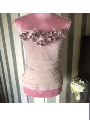 Ljusrosa axelbandslös topp - Supersöt ljusrosa axelbandslös topp med volangdetaljer och stora tygblommor upptill. Klänningen har en tight passform och är gjord i ett tunt, mjukt material som ger en snygg drapering. Perfekt för dig som vill sticka ut med en romantisk look.