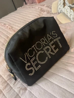 Svart necessär Victoria's Secret - Snygg svart necessär från Victoria's Secret med glittrande stenar som bildar loggan på framsidan. Väskan har en mjuk, rektangulär form och dragkedja upptill. Perfekt för smink eller småprylar. Materialet är troligtvis nylon eller polyester.