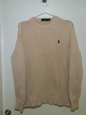 Beige stickad tröja Polo Ralph Lauren - Klassisk beige stickad tröja från Polo Ralph Lauren med rund halsringning och ribbade muddar. Tröjan har den ikoniska broderade loggan på bröstet och är tillverkad i ett mjukt material som passar perfekt till lager på lager.