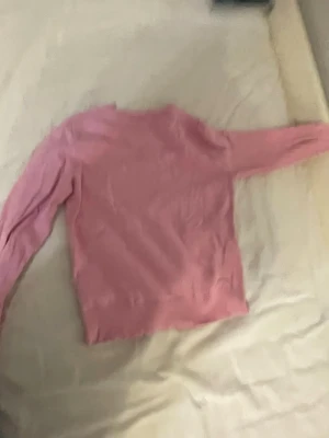 Rosa rundhalsad stickad tröja - Mjuk rosa stickad tröja med rund hals och långa ärmar. Tröjan har ribbade muddar vid ärmslut och nederkant för en clean look. Perfekt att bära över en t-shirt eller skjorta. Materialet ser ut att vara bomull eller en bomullsblandning. Size 140 cm