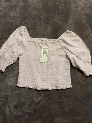 Ljuslila smockad topp KappAhl - Supersöt ljuslila topp från KappAhl med smockad kropp och puffiga trekvartsärmar. Toppen har en lätt volangkant nedtill och är sydd i ett mjukt bomullstyg med diskret prickigt mönster. Perfekt för dig som gillar romantisk och trendig stil.