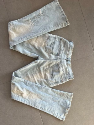  Broderade vintage jeans med pärlor  - Säljer ett par ljusblå bootcut/raka jeans med snygga broderier med pärlor och unika bakfickor med dekorativa sömmar. Står ingen storlek med för referens är jag 165 cm och väger ca 53 kg. Några pärlor har lossnat på vänster ben.