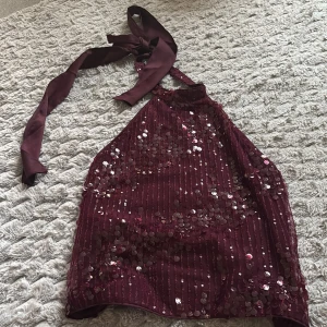 Vinröd paljett-topp med knyt - Snygg vinröd topp med paljetter och knyt i nacken. Toppen har halterneck-modell och är croppad, perfekt för att sticka ut. Materialet känns som polyester och satin, vilket ger ett glansigt intryck. Passar dig som vill ha något extra till outfiten.
